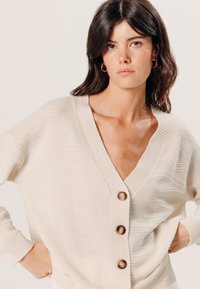 Hellbeige Strickjacke mit V-Ausschnitt, strukturiertem Stoff und fünf Holzknöpfen auf der Vorderseite. Mit gerippten Bündchen und Saum.