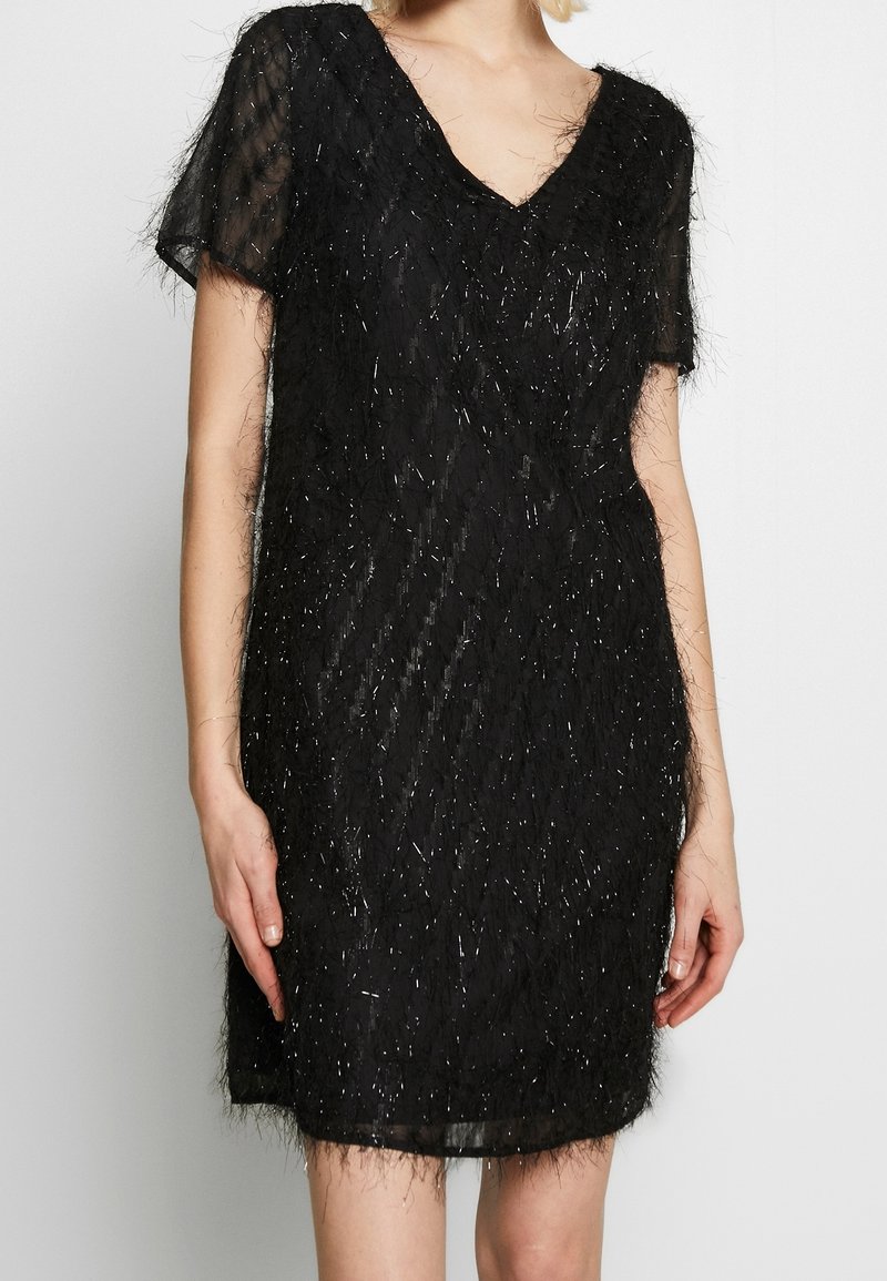 Femme portant une robe noire à manches courtes avec un col en V, en tissu texturé et duveteux avec des fils subtilement brillants.