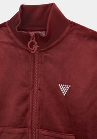 Veste zippée bordeaux avec un col haut, fabriquée en matériau doux. Présente un design en strass triangulaire sur la poitrine et une fermeture éclair.