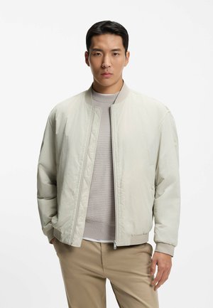 Uomo che indossa una giacca bomber beige chiaro sopra un maglione grigio e pantaloni kaki, in piedi contro uno sfondo bianco semplice.