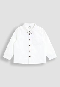 Camicia bianca a maniche lunghe con colletto, caratterizzata da bottoni marroni lungo la parte anteriore e una piccola tasca sul petto. Realizzata in un tessuto leggero.