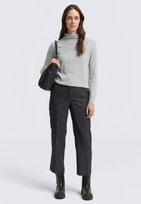 Grijze coltrui gecombineerd met donkergeltexteerde broek en zwarte enkellaarsjes. Model draagt een zwarte schoudertas. Eenvoudig, modern ontwerp.