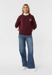 Vrouw staat met een wit shirt onder een bordeauxrode sweatshirt, blauwe uitlopende jeans en witte sneakers tegen een witte achtergrond.