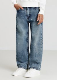 Jeans large en denim bleu clair avec des accents en cristal. Comprend des poches avant et une finition légèrement déchirée. Associés à des chaussures blanches.