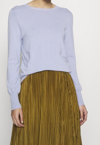 Woman wearing a light purple textured long-sleeve top tucked into a mustard yellow pleated skirt, standing against a plain background.