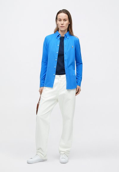 Modrá košeľa na gombíky z ľahkej látky, s dlhými rukávmi a golierom, doplnená bielymi nohavicami so širokými nohavicami a bielymi teniskami.