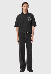 Zwarte oversized t-shirt met grafisch ontwerp aan de voorkant, gecombineerd met losse zwarte jeans. Model staat en draagt zwarte schoenen en ringen.