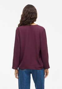 Frau mit langen, welligen braunen Haaren, die ein strukturiertes langärmliges lila Oberteil und blaue Jeans trägt und mit dem Rücken zu einem weißen Hintergrund steht.