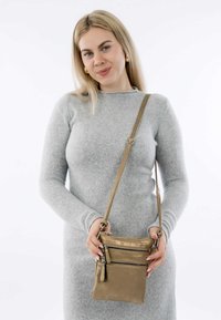 Borsa a tracolla metallica dorata con numerosi scomparti con zip e finitura texturizzata, tenuta da una persona in un maglione grigio chiaro.