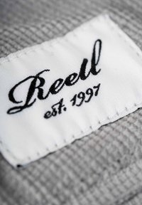 White fabric tag with "Reell" in black cursive, established year "1997" beneath, stitched onto a textured gray material.