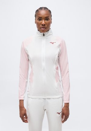 Γυναίκα που στέκεται αντικρίζοντας μπροστά, φορώντας λευκό και ανοιχτό ροζ αθλητικό μπουφάν με φερμουάρ και αντίστοιχο παντελόνι με λογότυπα.