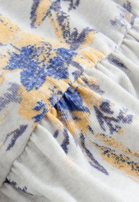 Gros plan d'un tissu blanc doux avec un imprimé floral texturé bleu et jaune et un détail de couture froncée.