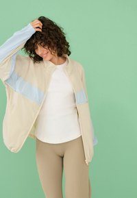 Jeune femme aux cheveux bouclés portant une veste beige et bleue sur une chemise blanche et un pantalon beige, souriante et touchant ses cheveux devant un fond vert.