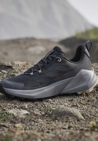 Chaussure de trail running Adidas Terrex noire et grise sur un sol rocailleux avec semelle extérieure texturée antidérapante et languette au talon.