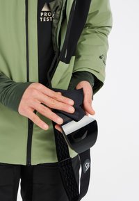 Una persona con una giacca verde e nera tiene un panno di pulizia nero contro un paio di occhiali da sole neri con lenti lucide e cinturino.