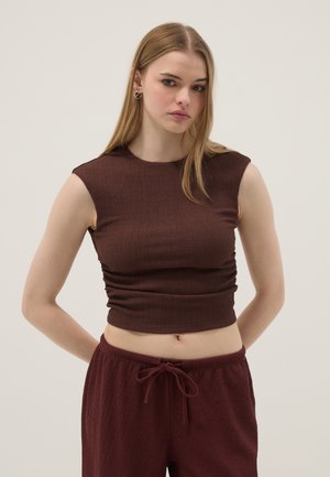 Jeune femme aux longs cheveux blonds portant un haut court brun sans manches et un pantalon assorti à cordon, debout les mains derrière le dos.