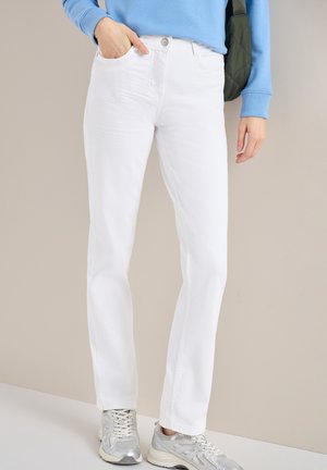 Osoba ubrana w białe spodnie o prostych nogawkach, srebrne trampki, jasnoniebieską bluzę i ciemnozieloną torbę na ramię, stojąca na tle beżowej ściany.