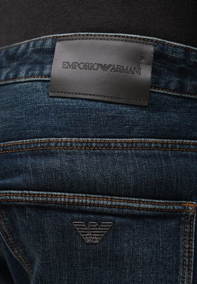 Sötétkék farmernadrág narancssárga varrással, az Emporio Armani fekete bőrcímkéjével és fémből készült saslogójával a hátsó zsebében.