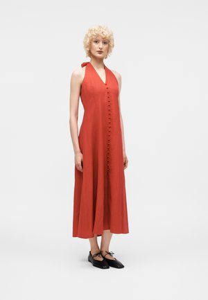 Kobieta z kręconymi blond włosami, ubrana w bezrękawnikową, rdzawej czerwieni sukienkę midi z zakrytymi z przodu guzikami i czarne baleriny, stojąca na białym tle.