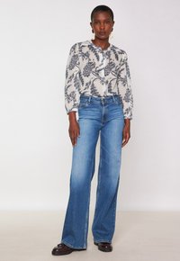 Donna che indossa jeans larghi blu, una blusa bianca con stampe floreali blu navy e di pavone, mocassini marroni e grandi orecchini a goccia di perle.