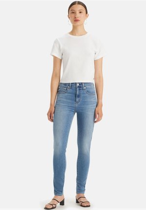 Camiseta de algodón blanca, de mangas cortas, combinada con jeans ajustados de un azul claro, con un desgastado sutil. Llevada con sandalias de tiras negras.