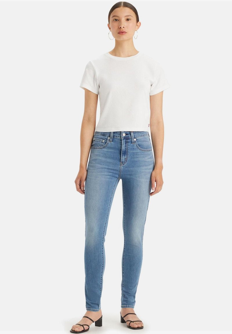 Camiseta de algodón blanca, de mangas cortas, combinada con jeans ajustados de un azul claro, con un desgastado sutil. Llevada con sandalias de tiras negras.