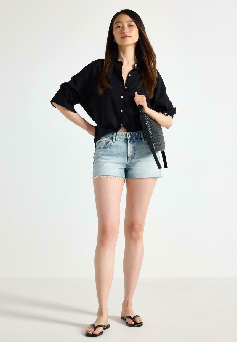 Svart knapp-up skjorta, denimshorts och svarta flip-flops. Skjortan har en avslappnad passform och uppvikta ärmar; shortsen har en fransad nederkant.