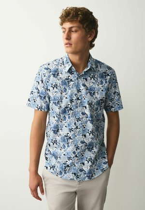 Young man looking left wearing short-sleeve button-up shirt with blue floral pattern and light-colored pants against plain background.