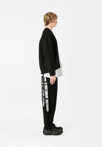 Suéter negro de punto texturizado sobre una camisa blanca, combinado con pantalones negros que presentan un estampado gráfico en el lateral y zapatos en negro de suela gruesa.