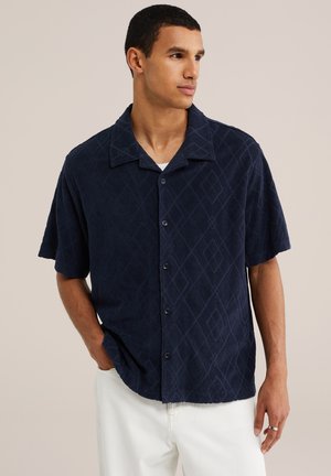 Homme portant une chemise à manches courtes bleu foncé avec un motif subtil en losanges et un pantalon blanc, debout avec une main dans la poche.