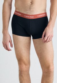 Črne bombažne bokserice s rdečim pasom z logotipom Calvin Klein. Imajo prileganje in mehko teksturo, brez vidnih vzorcev.