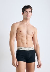 Oben ohne Mann mit kurzen dunklen Haaren, der schwarze Calvin Klein Boxershorts mit weißem Bund trägt, steht vor einem schlichten hellen Hintergrund.