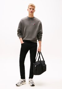 Siva majica z rebrastimi manšetami, črne kavbojke in sivi športni copati. Model drži črno športno torbo z zadrgo in logotipom.