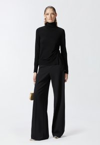 Pull à col roulé noir avec fronces sur les côtés, associé à un pantalon noir à jambes larges. Le mannequin tient un petit sac métallisé doré.