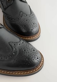 Čierne kožené brogues s detailným perforovaním, hnedým prešívaním pozdĺž podrážky a hladkým lesklým povrchom na svetlom pozadí.