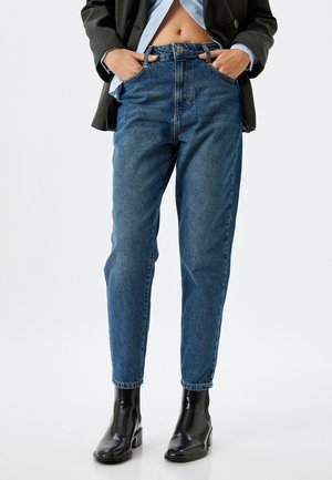 Jean en denim bleu taille haute avec une coupe décontractée, design à cinq poches et fermeture à bouton à l'avant, associés à des bottines noires.