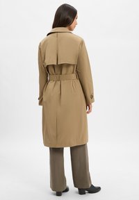 BeigerTrenchcoat mit tailliertem Gürtel, Schulterklappen und langen Ärmeln. Getragen über olivgrünen Hosen und schwarzen Stiefeletten mit Absatz.