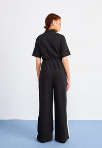 Svart jumpsuit med brede ben, korte ermer og elastisk liv. Har hvite side-striper. Materialet ser mykt og strukturert ut.