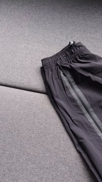 Calças de treino pretas com cintura elástica, bolsos laterais com fecho éclair e riscas cinzentas ao longo de uma textura lisa sobre uma superfície cinza.