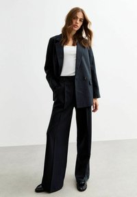 Completo blu a righe verticali con giacca doppiopetto e pantaloni a gamba larga, abbinato a una maglietta bianca e scarpe nere. Tessuto di superficie liscia.