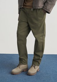 Άτομο που φορά χακί παντελόνι και μπεζ δερμάτινα μποτάκια με κορδόνια, στέκεται σε μπλε χαλί, με τα χέρια στις τσέπες, με καφέ σακάκι μερικώς ορατό.