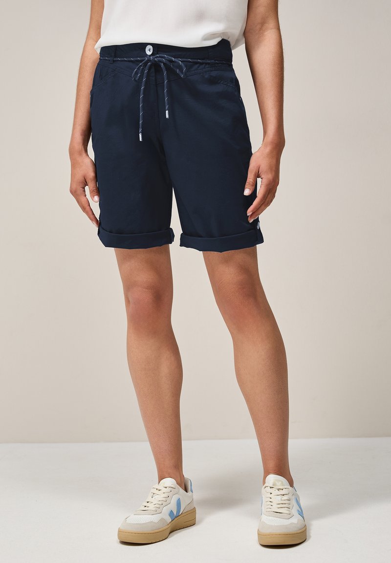 Marineblauwe shorts met een verstelbare tailleband, zijzakken en opgerolde mouwen, gemaakt van lichtgewicht stof. Gepaard met witte sneakers.