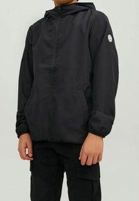 Černá bundu s kapucí z lehké tkaniny. Má přední zip, dvě boční kapsy a logo na levém rukávu.