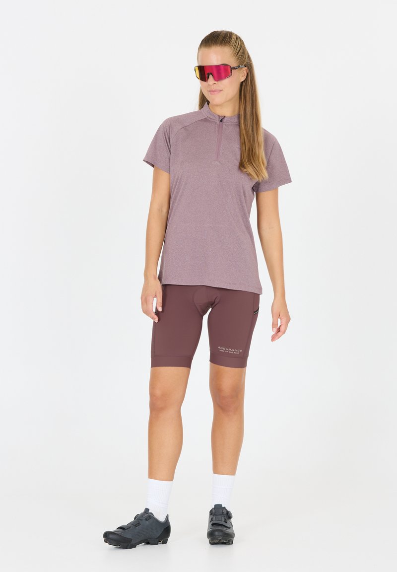 Maillot de cyclisme violet à manches courtes avec zip, short rembourré marron, chaussures de cyclisme noires, et lunettes de soleil réfléchissantes roses. Tissu lisse et respirant.