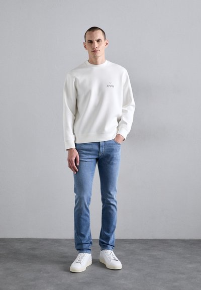 Sweatshirt alb cu guler rotund și un logo subtil, asortat cu blugi deschis la culoare și teniși albi, pe un fundal neutru.