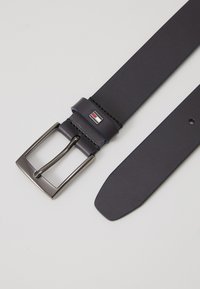 Ceinture en cuir bleu foncé avec une boucle rectangulaire en argent. Elle présente une boucle en cuir cousue et un petit accent de logo. Texture lisse sur toute la surface.