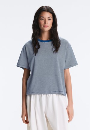 Frau mit langen dunklen Haaren, die ein blau-weiß dünn gestreiftes kurzärmeliges Hemd und lockere weiße Hosen trägt, steht vor einem einfarbigen Hintergrund.