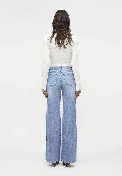 Žena s vlnitými vlasmi, oblečená v priliehavom bielom tričku s dlhým rukávom, vysokým pásom širokých svetlomodrých rifle a čiernymi opätkami, stojaca otočená chrbtom.