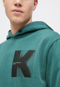 Hanorac teal cu un logo grafic negru în formă de "K" și accente liniare. Materialul pare moale, cu o finisare texturată. Include o glugă cu șnur.