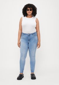 Light blue skinny jeans with a high waist, paired with a white sleeveless top and black slip-on shoes. Curly hair and sunglasses.